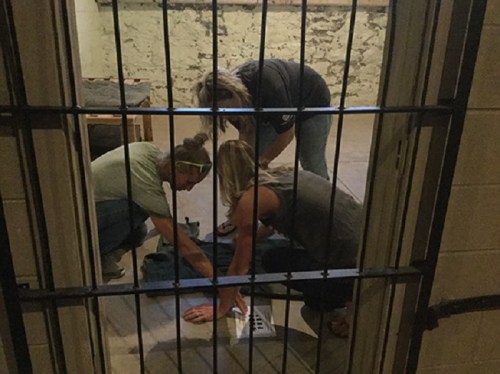 Three people crouch on the floor, examining an object in a dimly lit room behind bars.