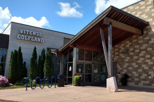 Entrance of Witense Golfland with a wooden awning, bike rack, and landscaped greenery under a blue sky.