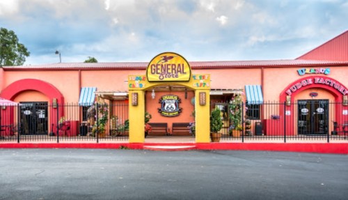 Colorful storefront of a general store with a fudge factory, featuring Route 66 signage and decorative flowers.