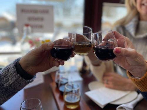 Three people clink glasses of red wine in a cozy setting, with a tasting flight of drinks in the background.