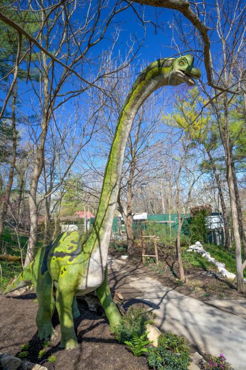 A large green dinosaur statue stands among trees in a park, with a clear blue sky in the background.