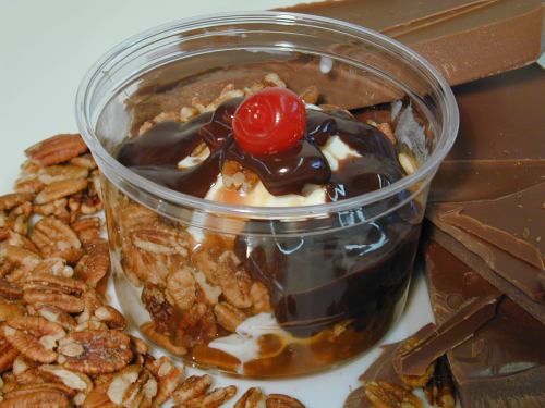 A dessert cup with layers of ice cream, chocolate sauce, nuts, and a cherry on top, surrounded by chocolate bars and pecans.