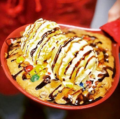 A warm cookie dessert topped with two scoops of ice cream, chocolate syrup, and colorful candy pieces in a red bowl.