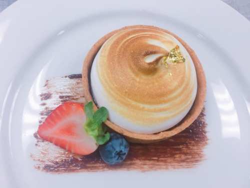 A beautifully plated dessert featuring a golden-brown meringue atop a tart shell, garnished with a strawberry and blueberry.