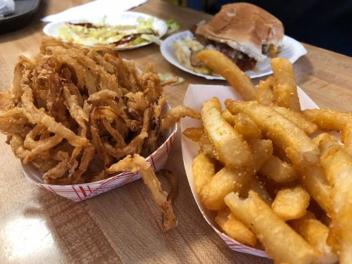 A close-up of crispy fried onions and golden French fries on a wooden table, with a burger and lettuce in the background.