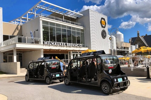 Two electric shuttles parked outside Milwaukee Brewing Co., with a modern building and outdoor seating in the background.