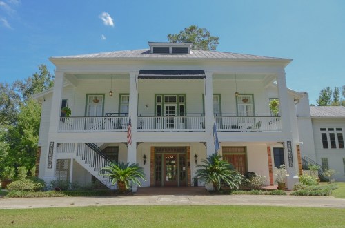 A large white two-story house with a porch, stairs, and greenery, set against a clear blue sky.