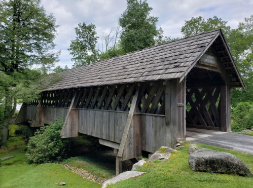 A wooden covered bridge with a sloped roof, surrounded by greenery and rocks under a cloudy sky.