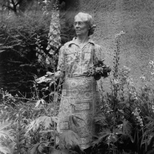 A woman in a patterned dress stands among tall flowers, holding a bouquet in a garden setting.