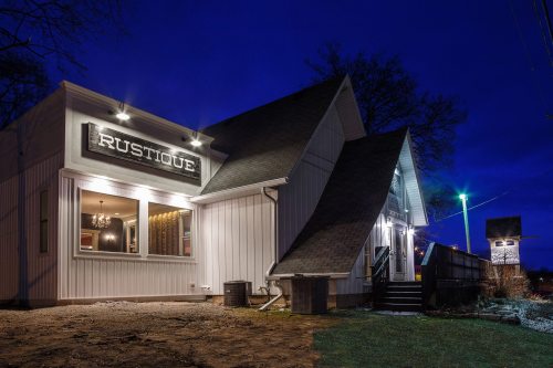 A modern building with a sign reading "Rustique," illuminated at night, featuring a sloped roof and outdoor lighting.