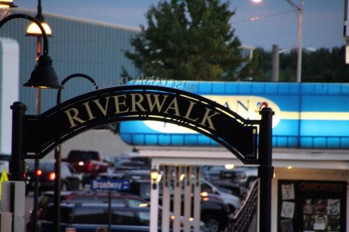 Sign reading "RIVERWALK" with a blue building and parked cars in the background during twilight.