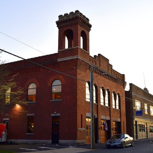 Historic brick building with arched windows and a tower, illuminated by evening light, parked car in front.