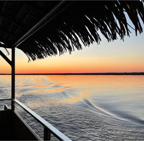 A serene sunset over calm waters, viewed from a thatched-roof deck, with gentle ripples reflecting the colors of the sky.