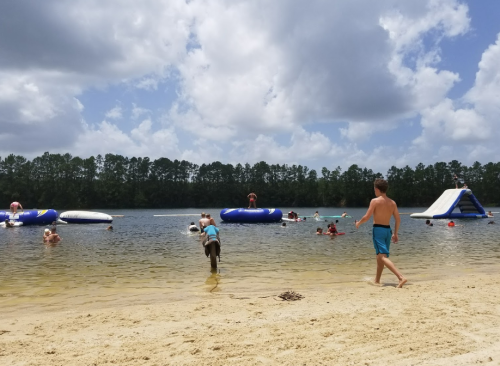 A sunny beach scene with people swimming and playing on inflatable structures in the water, surrounded by trees.
