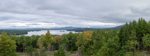 A panoramic view of a serene lake surrounded by lush green trees and distant mountains under a cloudy sky.