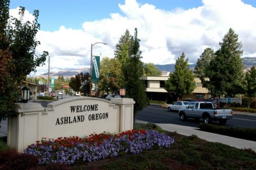 Welcome sign for Ashland, Oregon, with flowers and trees lining the street under a partly cloudy sky.