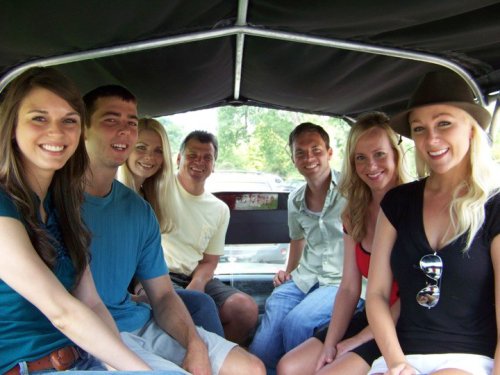 A group of eight smiling friends sitting together in a vehicle, enjoying a sunny day outdoors.