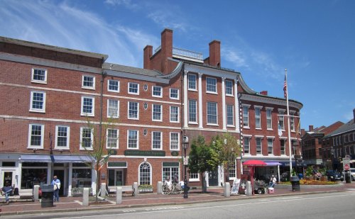 Historic brick building with large windows, trees, and outdoor seating, set against a clear blue sky.