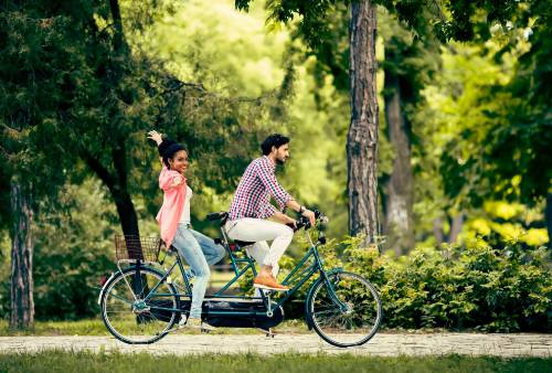 A man and woman ride a tandem bicycle through a lush green park, enjoying a sunny day together.
