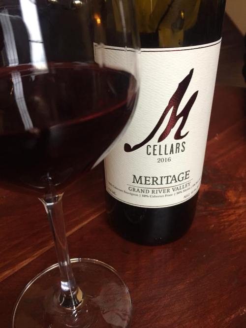 A glass of red wine beside a bottle labeled "M Cellars Meritage 2016" on a wooden table.