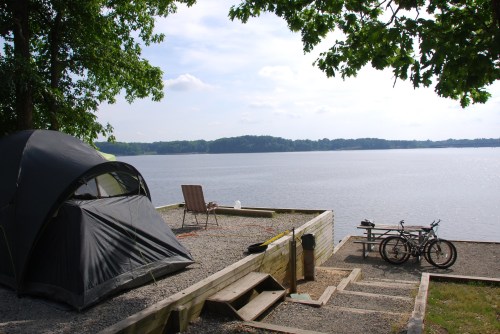 A tent and two bicycles by a calm lake, surrounded by trees and a gravel area with a chair.