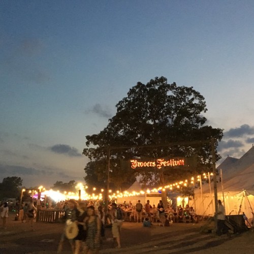 A festival entrance sign illuminated by string lights at dusk, with people walking around and tents in the background.