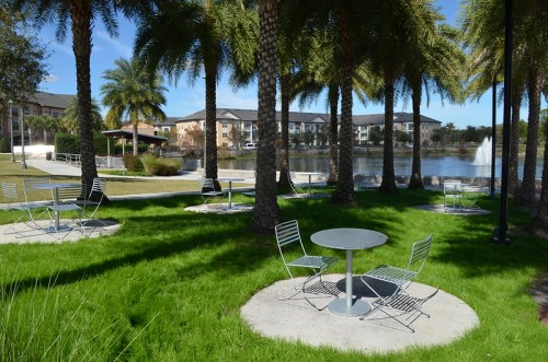 A serene park scene with palm trees, green grass, and metal tables and chairs near a pond and fountain.
