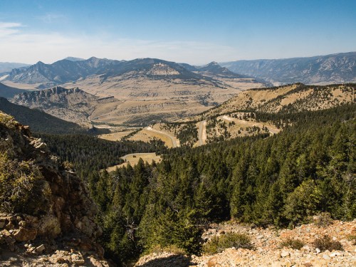 A panoramic view of rugged mountains and valleys, with dense forests and winding dirt roads under a clear blue sky.