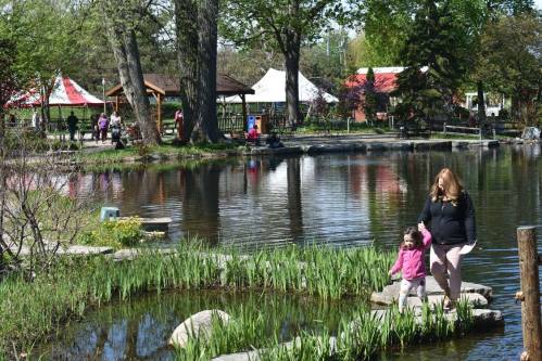 A woman and a child walk along a rocky path by a pond, surrounded by greenery and festival tents in the background.