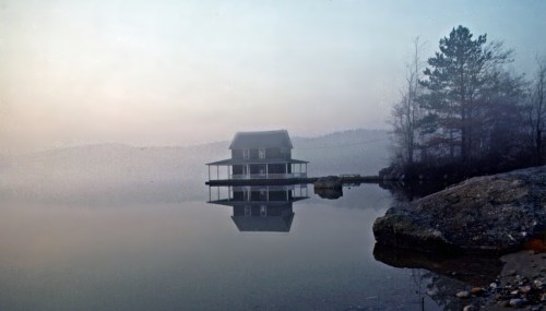 A misty lake scene featuring a house on stilts, reflecting in the calm water, surrounded by trees and rocks.
