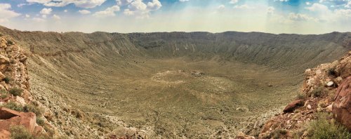 A panoramic view of a large, dry crater surrounded by rocky terrain and sparse vegetation under a blue sky.