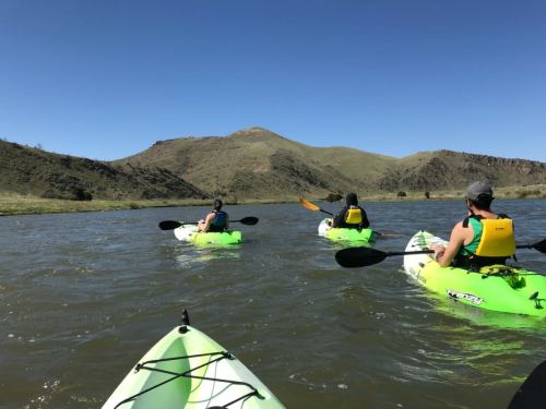 Three kayakers in green kayaks paddle on a calm river, surrounded by rolling hills under a clear blue sky.