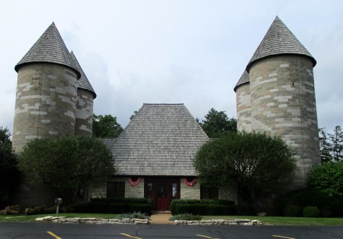 A stone building with two tall, conical towers and a sloped roof, surrounded by greenery and a paved parking area.