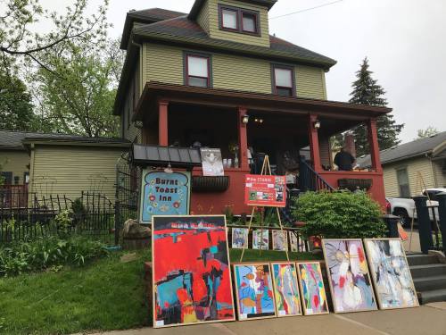 A colorful house with a sign for "Burnt Toast Inn" and art displayed on the lawn during an outdoor event.