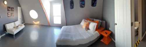 A modern bedroom with a bed, orange accents, a sofa, and a bathroom door, featuring unique circular windows.