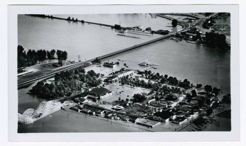 Aerial view of a flooded area with buildings, trees, and a bridge partially submerged in water. Black and white photo.