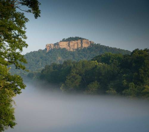 A misty landscape featuring a prominent rocky hill surrounded by lush green trees and a serene atmosphere.