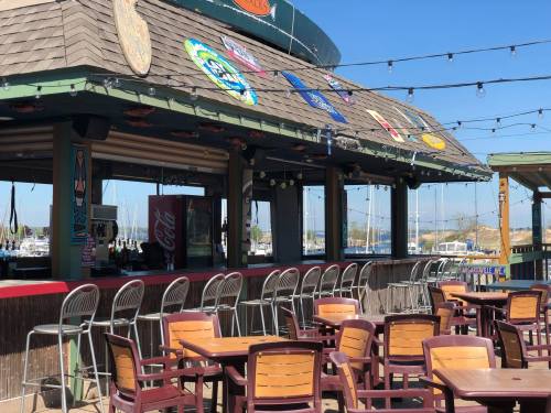 Outdoor bar with empty tables, colorful decorations, and a view of boats in the marina under a clear blue sky.