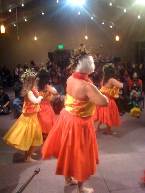 A group of dancers in colorful traditional attire perform on stage, with an audience watching in the background.