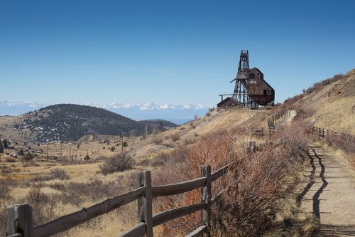A rustic building on a hillside with mountains in the background and a dirt path lined by a wooden fence.