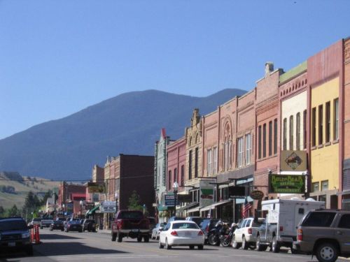A charming small town street lined with colorful historic buildings and parked cars, set against a backdrop of mountains.