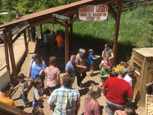 A busy outdoor area with families and children engaging in activities, surrounded by greenery and wooden structures.