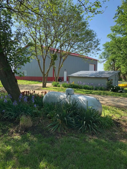 A propane tank surrounded by flowers and greenery, with a large barn and trees in the background on a sunny day.