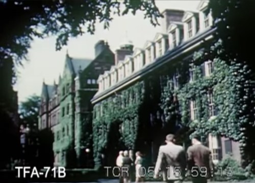 A historic building covered in ivy, with people walking along a path in a lush, green setting.