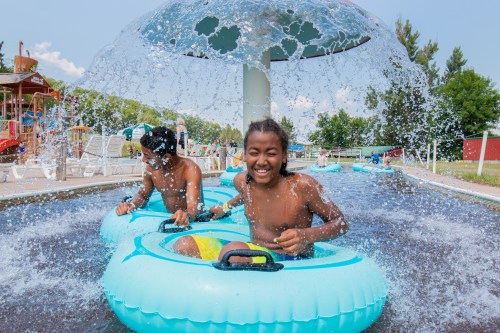 Two children in inflatable kayaks enjoy a splash pad, laughing as water sprays around them on a sunny day.