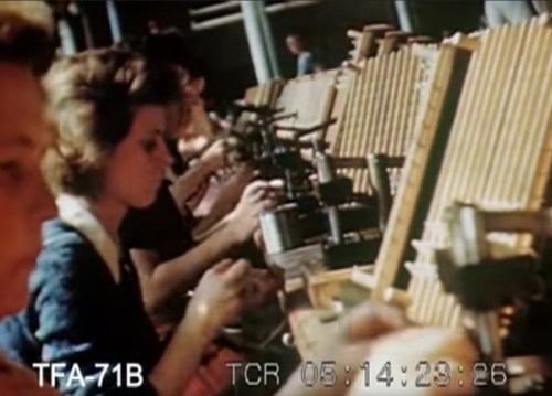 Women working at desks with machinery, focused on tasks in a factory setting. Colorful vintage footage.