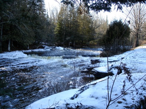 A serene winter scene featuring a flowing river surrounded by snow-covered banks and evergreen trees.