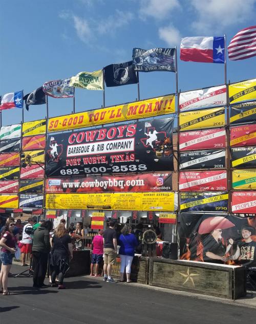 A busy barbecue stand with colorful flags and a large sign reading "Cowboys Barbeque & Rib Company" in Texas.