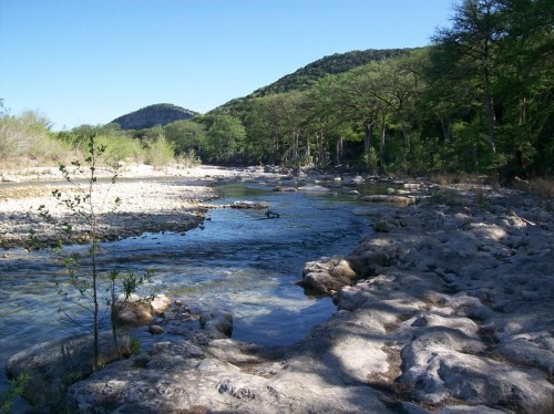 A serene river flows through rocky terrain, surrounded by lush trees and rolling hills under a clear blue sky.