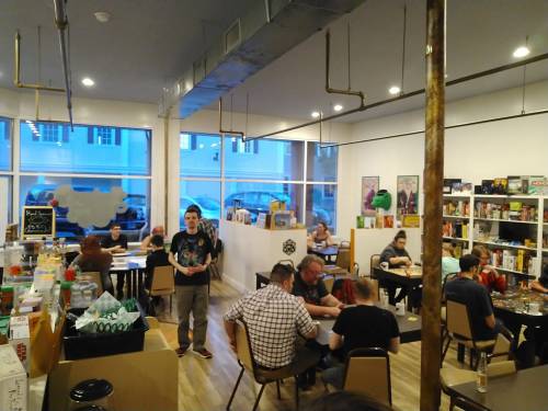 A cozy game café with people playing board games, shelves filled with games, and large windows letting in natural light.